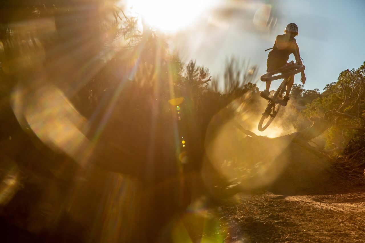 Im goldenen Licht der untergehenden Sonne fliegt ein Mountainbiker dynamisch über einen Trail-Absprung. Staub wirbelt auf, die Konturen verschwimmen im Lichtspiel – ein Moment voller Adrenalin, Freiheit und Naturverbundenheit. Der intensive Sonnenflare verstärkt die Dramatik der Szene und fängt die rohe Energie des Trails perfekt ein.