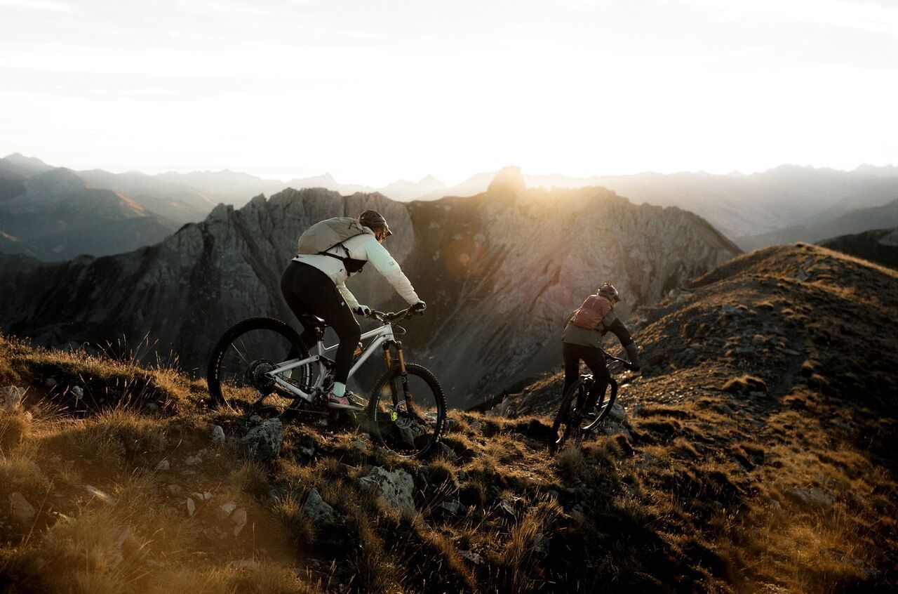 Zwei Mountainbiker fahren bei Sonnenuntergang auf einem schmalen, steinigen Berggrat, umgeben von beeindruckender alpiner Landschaft.
