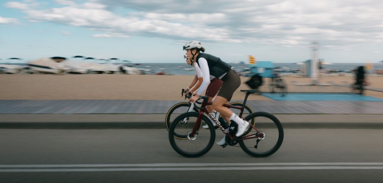 Zwei Radfahrerinnen fahren mit Rennrädern entlang einer Straße am Strand. Im Hintergrund sind Sand, das Meer und ein bewölkter Himmel zu sehen, während die Bewegung der Fahrerinnen Dynamik und Geschwindigkeit vermittelt.