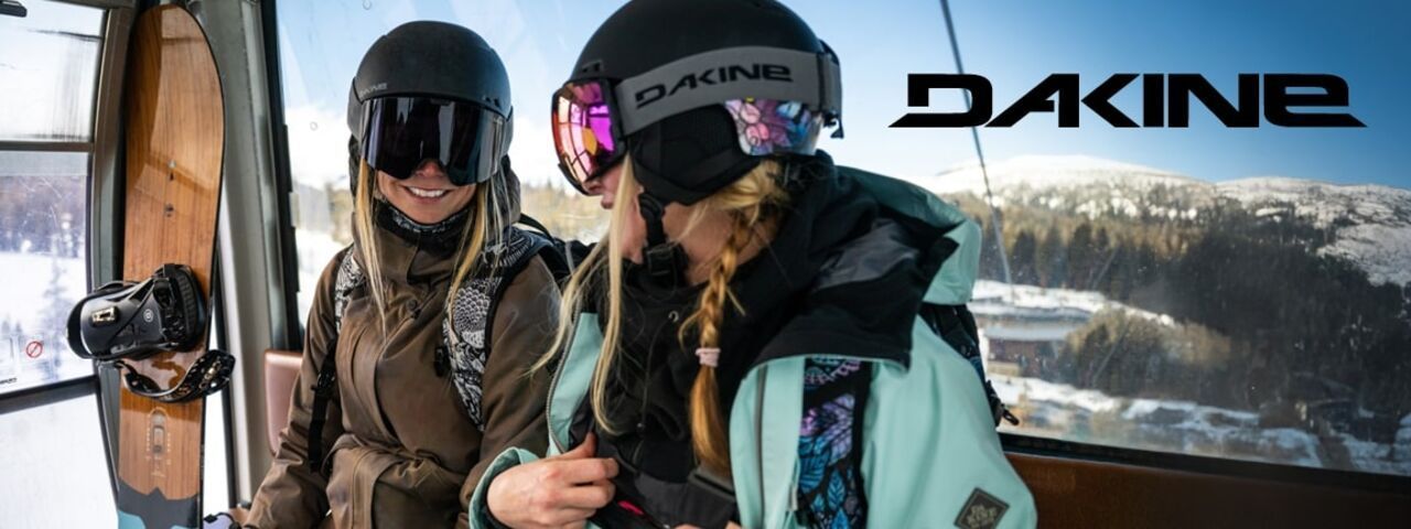 Zwei Snowboarderinnen in Dakine-Winterbekleidung in der Gondel mit Snowboard im Hintergrund – bereit für actionreiche Abfahrten im Schnee.