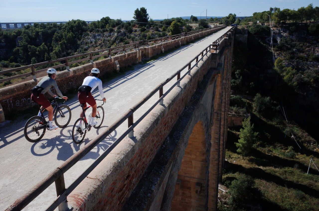 Zwei Rennradfahrer fahren über eine schmale Brücke durch eine sonnige Landschaft. Unter ihnen erstreckt sich ein tiefes Tal, während die Strecke geradeaus in die Ferne führt.