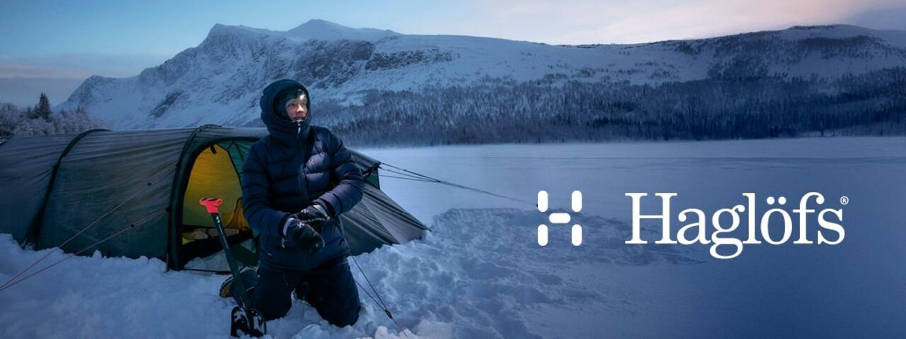 Winter-Camper mit Haglöfs-Ausrüstung vor Zelt in verschneiter Berglandschaft – funktionale Outdoor-Bekleidung für extreme Bedingungen.
