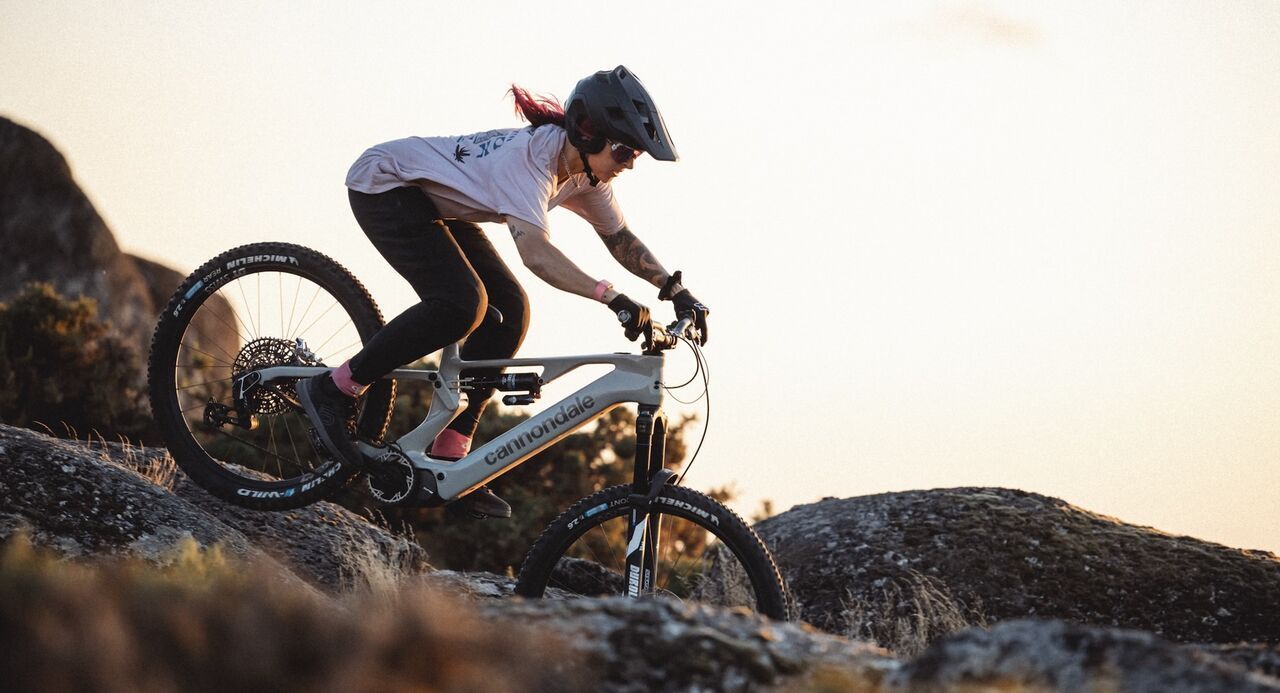Mountainbikerin auf einem Cannondale E-MTB fährt dynamisch über felsiges Terrain bei Sonnenuntergang – perfekte Kombination aus Technik, Style und Power.