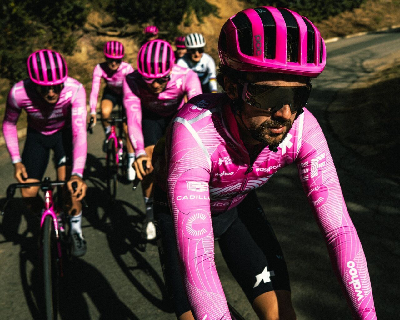 Eine Gruppe von Rennradfahrern in auffälligen pinken Trikots fährt dicht hintereinander eine Straße entlang. Im Vordergrund führt ein Fahrer das Feld an, während die anderen im Windschatten folgen.