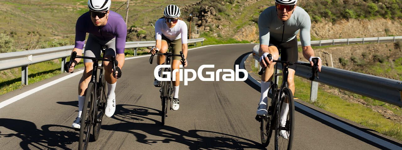 Drei Rennradfahrer fahren sportlich auf einer kurvigen Straße durch eine sonnige Landschaft. Sie tragen moderne Radbekleidung und Helme. In der Bildmitte steht der Schriftzug „GripGrab“, der die Szene als Markenmotiv kennzeichnet.
