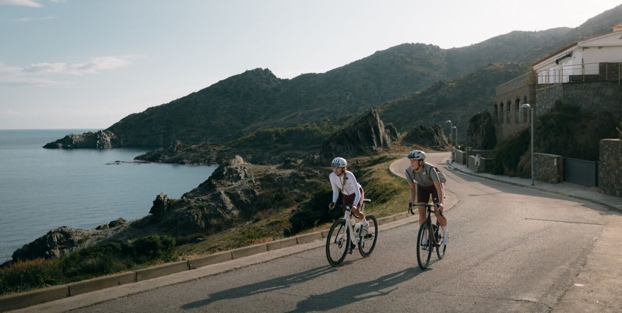 Zwei Rennradfahrerinnen fahren auf einer kurvigen Küstenstraße mit Blick auf das Meer und eine bergige Landschaft – ein Bild für sportliche Performance und Freiheit auf dem Rennrad.
