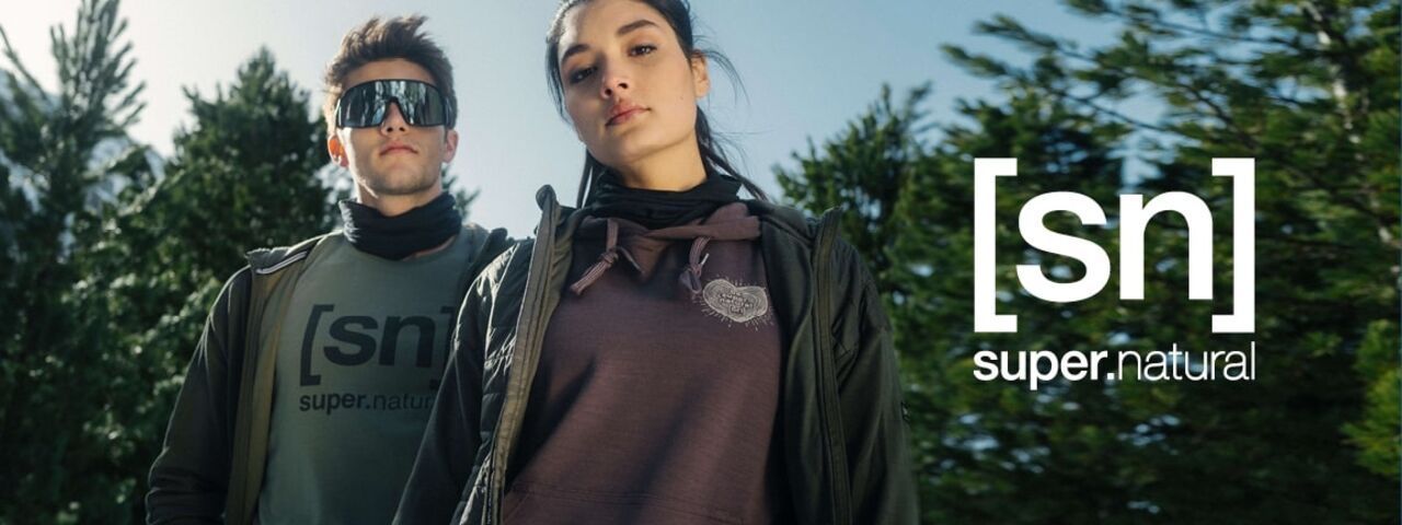 Das Bild zeigt zwei junge Erwachsene – eine Frau im Vordergrund und einen Mann dahinter – die sportliche Outdoor-Bekleidung der Marke super.natural tragen. Die Frau trägt einen dunkelvioletten Kapuzenpullover mit einem aufgestickten Herz-Logo sowie eine schwarze Jacke. Ihre dunklen Haare sind nach hinten gebunden. Der Mann trägt ein olivgrünes Shirt mit dem Logo [sn] super.natural, eine dunkle Jacke und eine auffällige, breite Sonnenbrille mit verspiegelten Gläsern. Beide Personen stehen vor einem Hintergrund aus grünen Nadelbäumen unter klarem, blauem Himmel. Rechts im Bild ist groß das weiße Logo der Marke eingeblendet: [sn] super.natural.