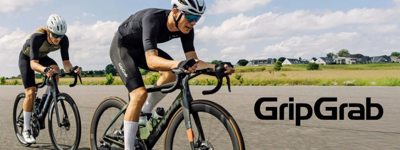 Zwei Radsportler im GripGrab-Outfit fahren dynamisch auf Rennrädern durch eine offene Landschaft mit Feldern und Häusern im Hintergrund. Sie tragen Helme, Sonnenbrillen und performancestarke Radsportbekleidung – ideal für intensives Training bei gutem Wetter.