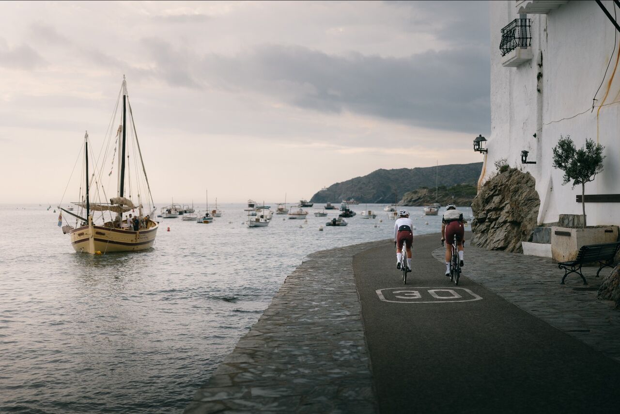 Zwei Rennradfahrer fahren entlang einer schmalen Küstenstraße direkt am Meer. Neben ihnen liegen zahlreiche Boote im Wasser, während im Hintergrund eine ruhige Bucht und hügelige Landschaft zu sehen sind.