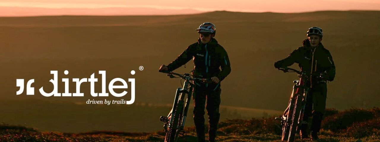 Zwei Mountainbiker mit Dirtlej-Bekleidung schieben ihre Bikes über einen Trail bei Sonnenuntergang – perfekt ausgestattet für Touren in anspruchsvollem Gelände.