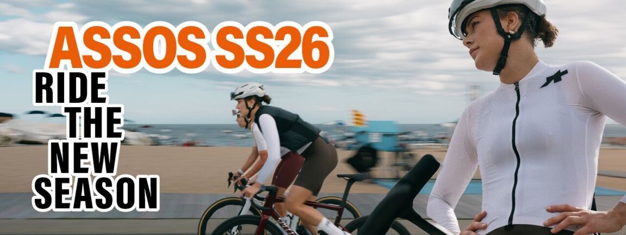 Zwei Rennradfahrerinnen fahren entlang einer Küstenpromenade, während im Vordergrund eine Athletin in weißem Trikot steht. Der Schriftzug „ASSOS SS26 – Ride the New Season“ unterstreicht den sportlichen und modernen Kampagnencharakter.