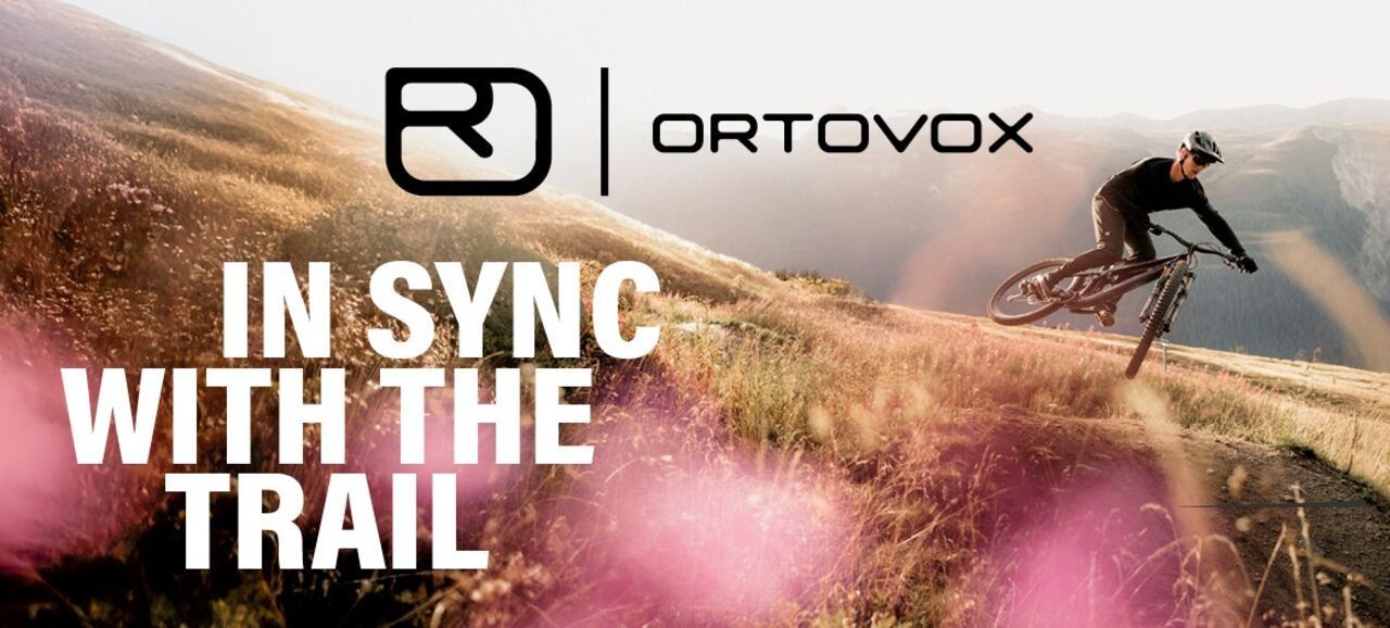 Mountainbiker fährt dynamisch einen Trail in alpiner Landschaft, begleitet vom Ortovox-Logo und dem Slogan „In Sync with the Trail“.