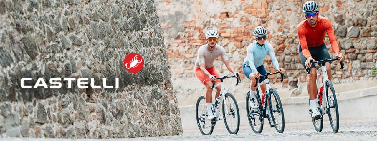 Drei Rennradfahrer fahren in sportlicher Kleidung durch eine enge, gepflasterte Straße entlang einer alten Steinmauer; links ist das Castelli-Logo eingeblendet.