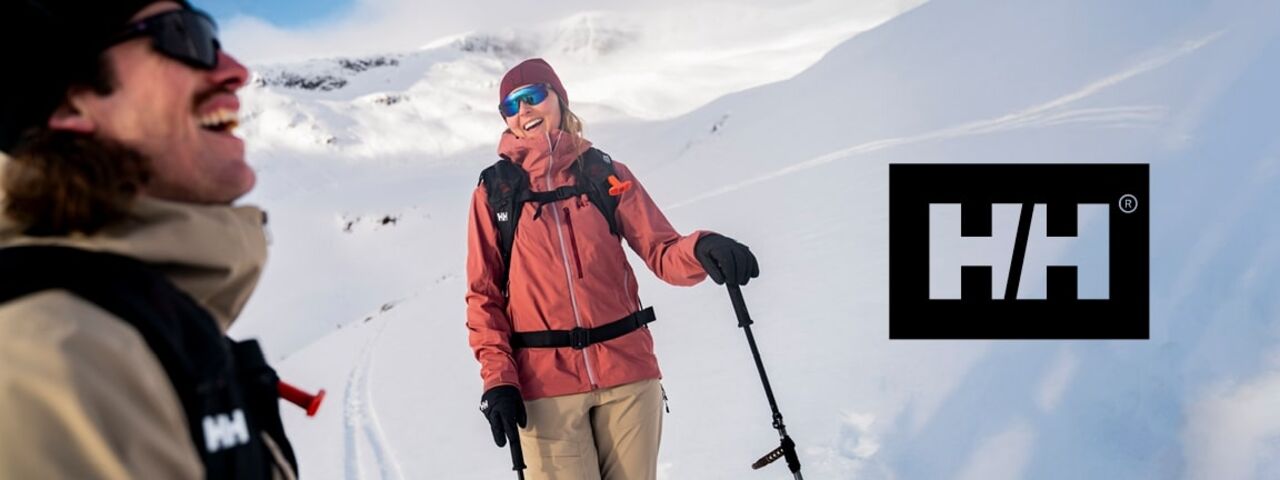 Zwei Skitourengeher in Helly-Hansen-Outfits genießen die Sonne im verschneiten Hochgebirge – perfekte Ausrüstung für alpine Abenteuer.