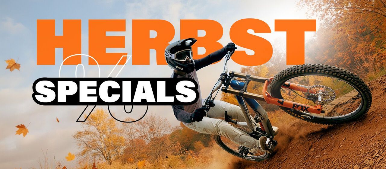 Mountainbike-Action im Herbst: Fahrer mit Helm und Schutzkleidung bei einem Sprung auf einem orangefarbenen Mountainbike vor herbstlicher Waldkulisse.