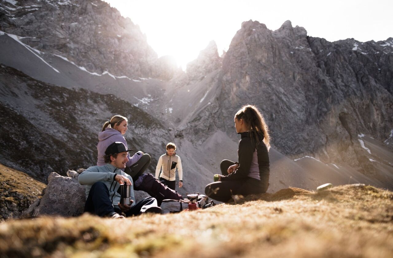 Inmitten einer beeindruckenden Berglandschaft genießen vier Freund:innen eine entspannte Pause beim Wandern. Umgeben von schroffen Felswänden und sanftem Sonnenlicht, sitzen sie gemütlich auf einer Wiese, teilen heißen Tee und gute Gespräche. Die Szene vermittelt Ruhe, Naturverbundenheit und das besondere Gemeinschaftsgefühl, das nur gemeinsame Outdoor-Abenteuer schaffen.