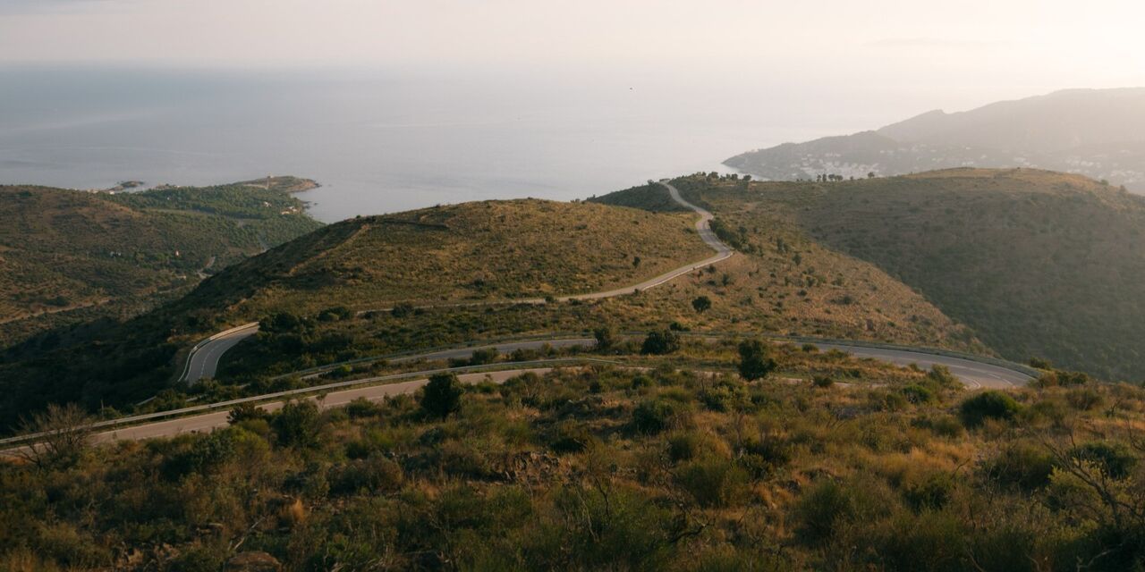 Das Bild zeigt eine hügelige Küstenlandschaft mit trockener Vegetation. Eine kurvige Straße schlängelt sich in Serpentinen durch die Hügel. Im Hintergrund liegt das ruhige Meer, rechts sind einige Gebäude entlang der Küste zu erkennen. Die warme Lichtstimmung wirkt wie spätnachmittags oder abends.