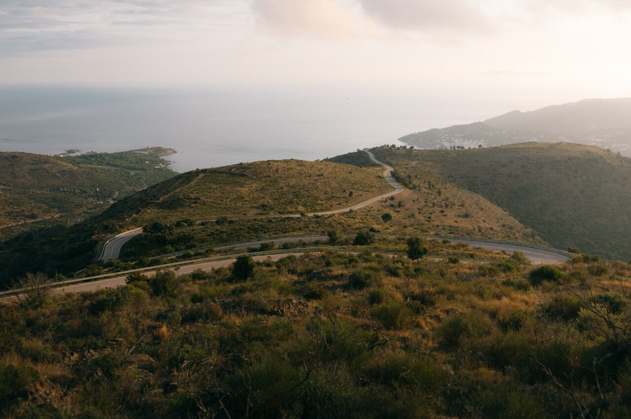 Das Bild zeigt eine hügelige Küstenlandschaft mit trockener Vegetation. Eine kurvige Straße schlängelt sich in Serpentinen durch die Hügel. Im Hintergrund liegt das ruhige Meer, rechts sind einige Gebäude entlang der Küste zu erkennen. Die warme Lichtstimmung wirkt wie spätnachmittags oder abends.