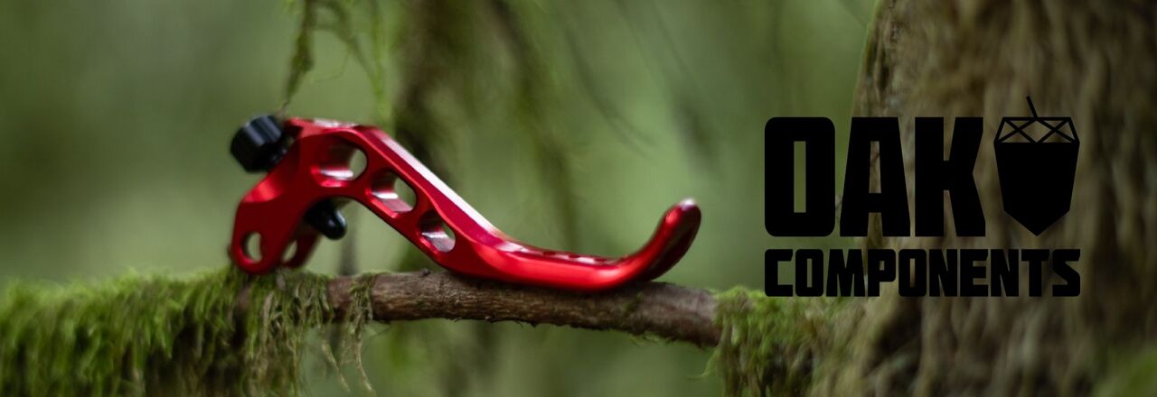 Detailaufnahme eines leichten, rot eloxierten Mountainbike-Bremshebels von OAK Components, inszeniert in natürlicher Waldumgebung.