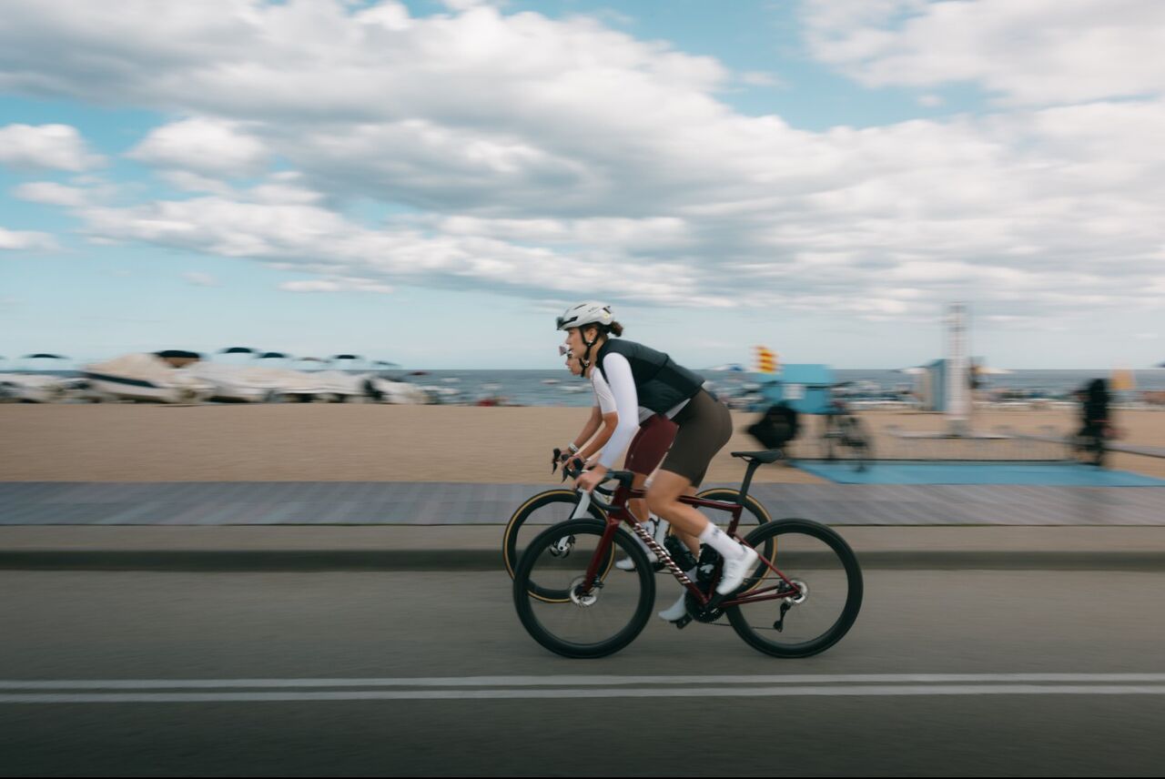 Zwei Radfahrerinnen fahren mit Rennrädern entlang einer Straße am Strand. Im Hintergrund sind Sand, das Meer und ein bewölkter Himmel zu sehen, während die Bewegung der Fahrerinnen Dynamik und Geschwindigkeit vermittelt.