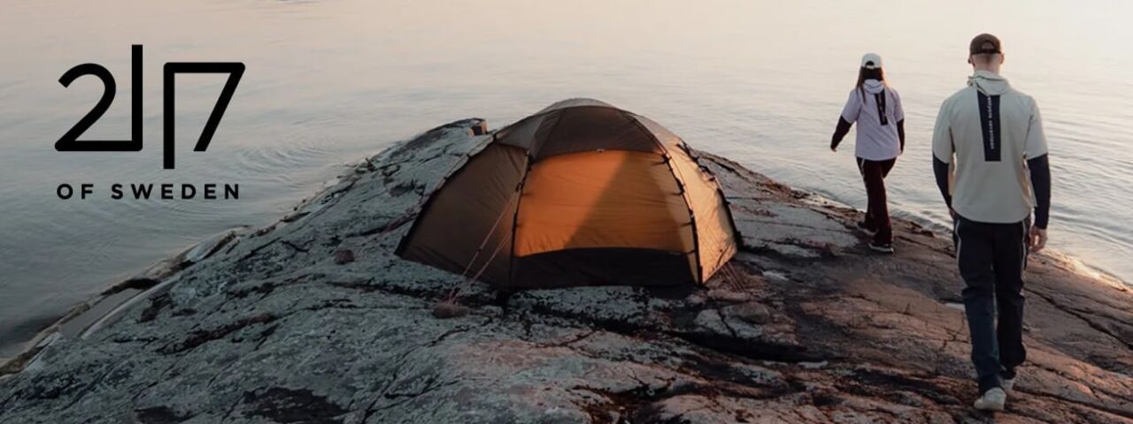 Zwei Personen in moderner Outdoor-Bekleidung von 2117 of Sweden auf einem felsigen Ufer mit Zelt, im Licht des Sonnenuntergangs – perfekter Start in ein Outdoor-Abenteuer am Wasser.