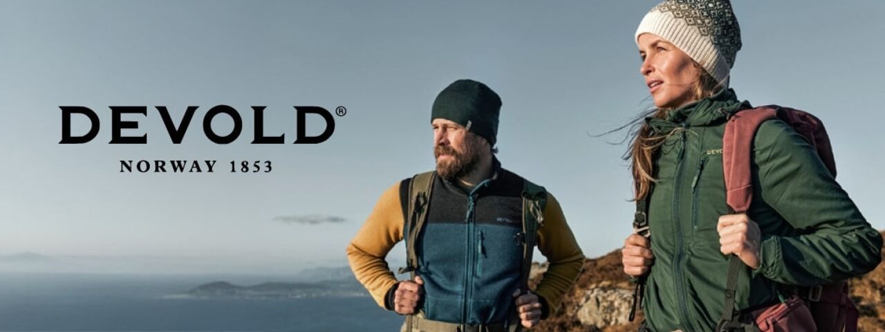 Zwei Wanderer in funktionaler DEVOLD-Outdoorkleidung genießen den Ausblick auf norwegische Fjorde – perfekt ausgerüstet für Abenteuer in der Natur.