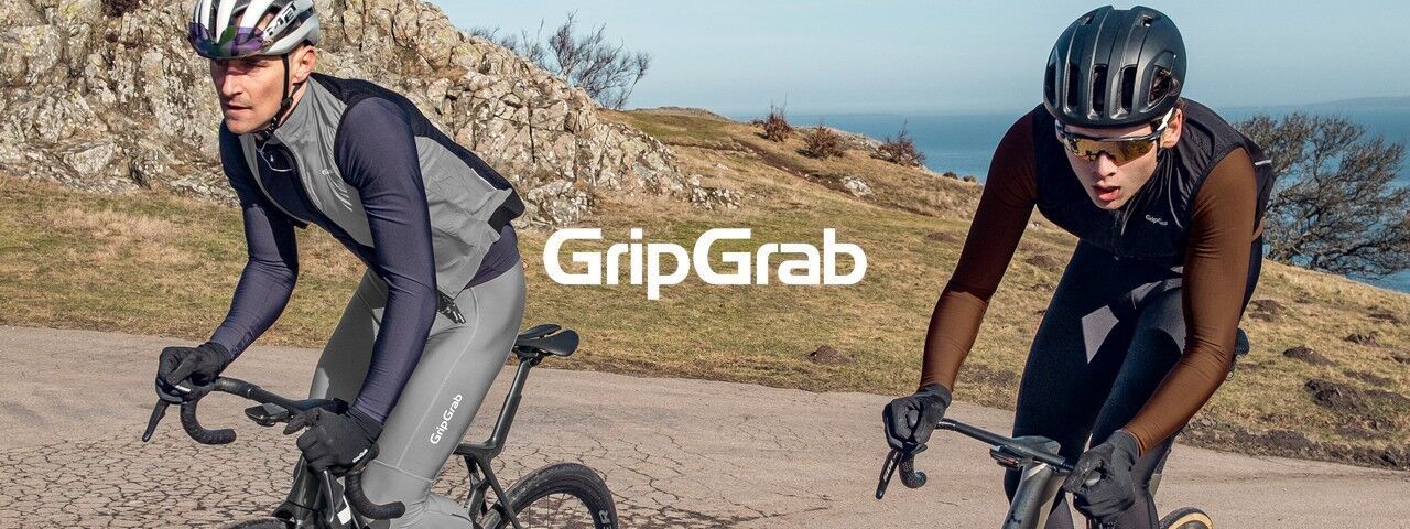 Zwei Rennradfahrer in sportlicher Fahrradbekleidung fahren auf einer Straße durch eine felsige Küstenlandschaft. In der Bildmitte steht das weiße GripGrab-Logo.