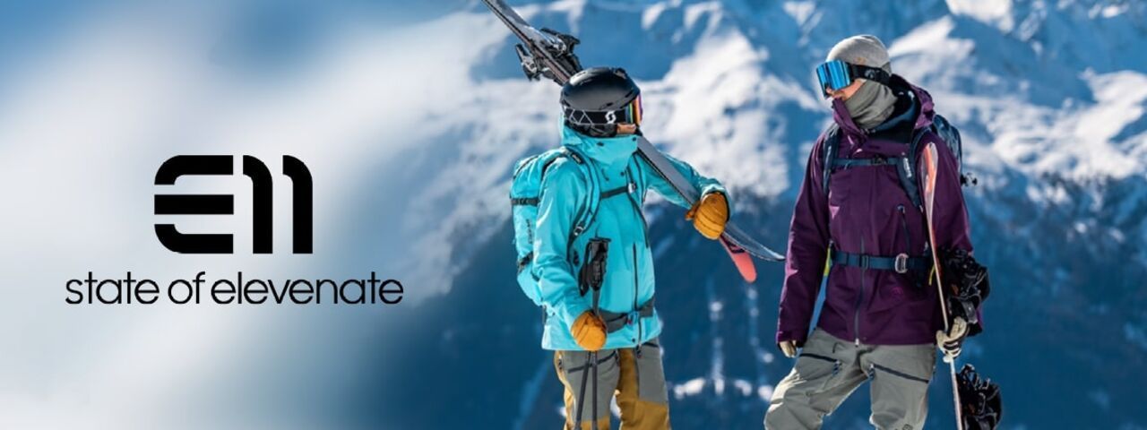 Stylische Skibekleidung im Schnee: Zwei Freerider im Gespräch, umgeben von verschneiten Berggipfeln – State of Elevenate vereint Funktionalität und Design.