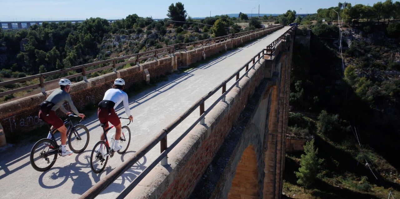 Zwei Rennradfahrer fahren über eine schmale Brücke durch eine sonnige Landschaft. Unter ihnen erstreckt sich ein tiefes Tal, während die Strecke geradeaus in die Ferne führt.